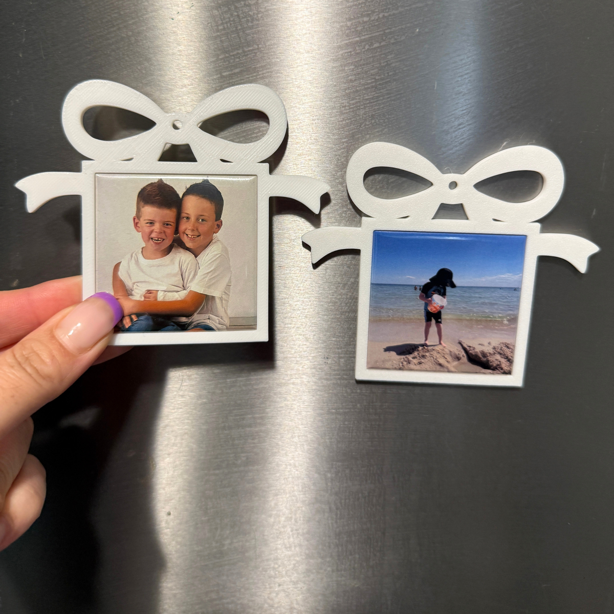 White present-shaped photo magnet ornaments displayed on a stainless steel fridge, showing two smiling boys and a beach photo